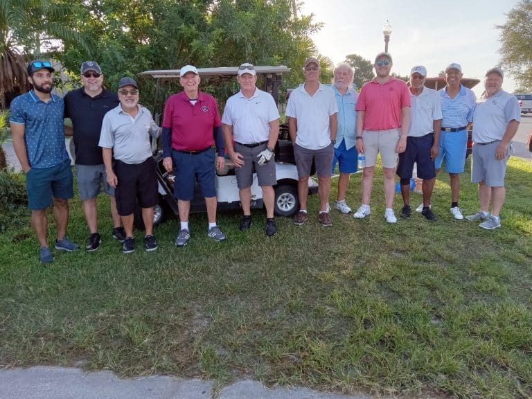 Group of men standing near a golf cart on a green lawn, some wearing hats and polo shirts.