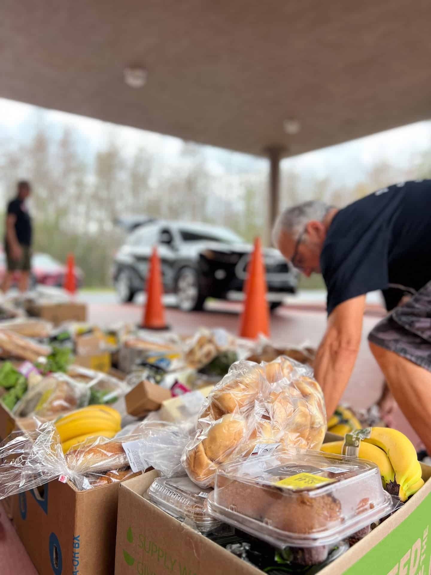 People sorting food donations outdoors. Cardboard boxes overflowing with items. Car and traffic cones in background.