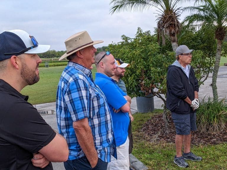 Group of men on a golf course, wearing hats, watching something off-camera.