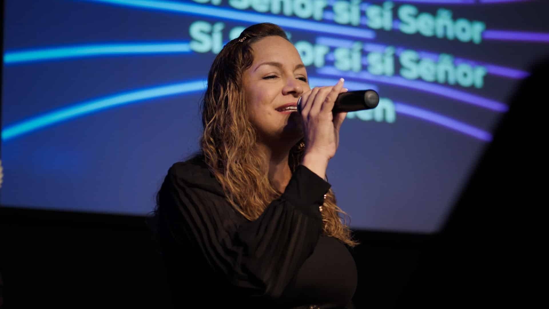 Woman singing into a microphone on stage, blue background with lyrics.