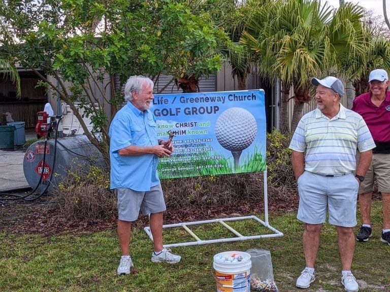 Men at a golf group event. One holds a trophy, others stand near a sign with a golf ball image.