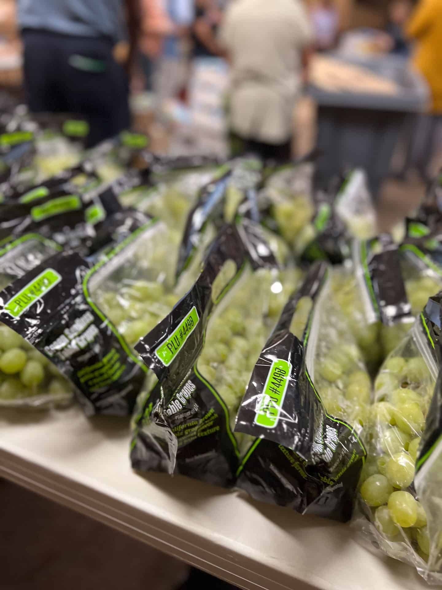 Bags of green grapes on a table with people in the background, likely a grocery store.