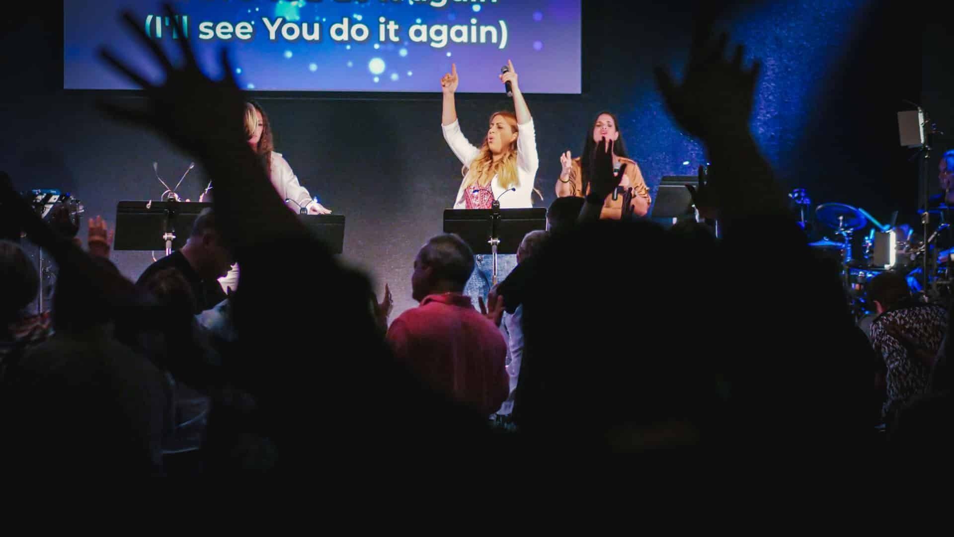 Worship band onstage with raised arms. Audience also has arms raised, lit in blue light. Lyrics displayed overhead.