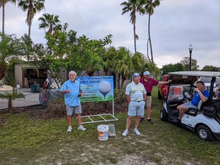 Men standing near a golf sign. Golf cart, trees, and buildings in background. Cloudy sky.