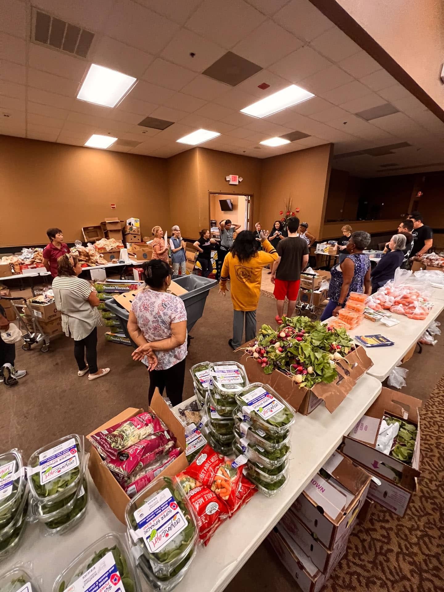 People at a food distribution event in a large room with tables of produce and goods.