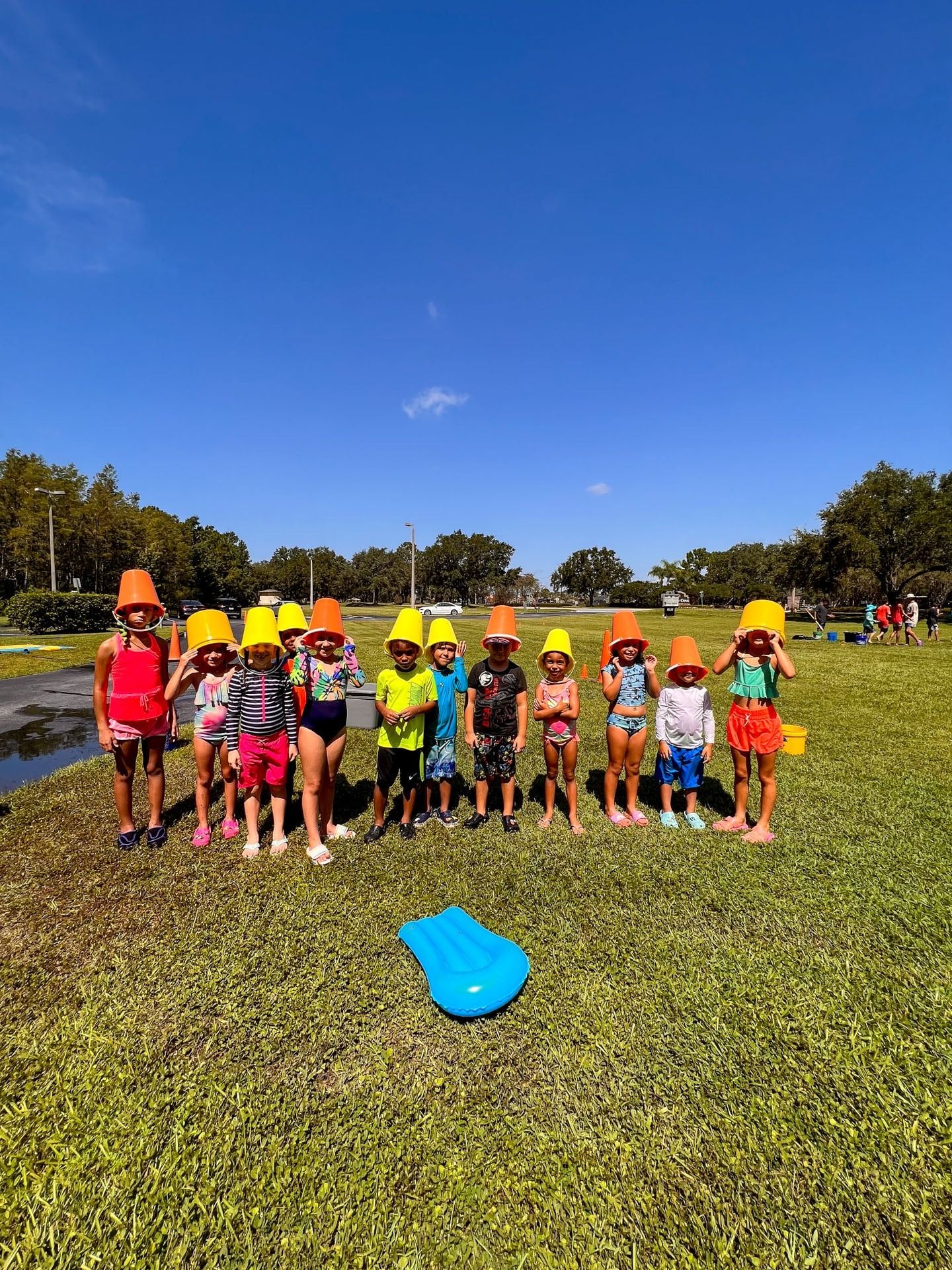 Children with orange buckets on their heads stand on grass under a blue sky.