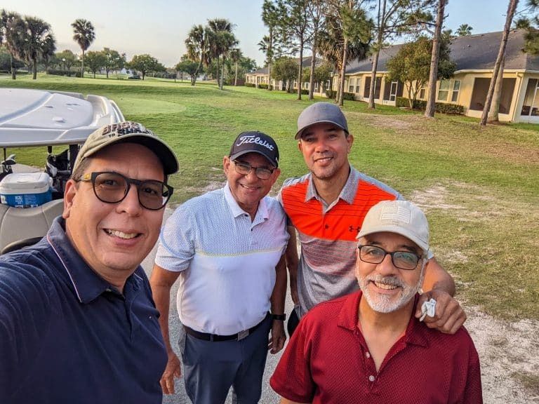 Four men smiling, posing for a selfie on a golf course. They stand near a golf cart on a sunny day.