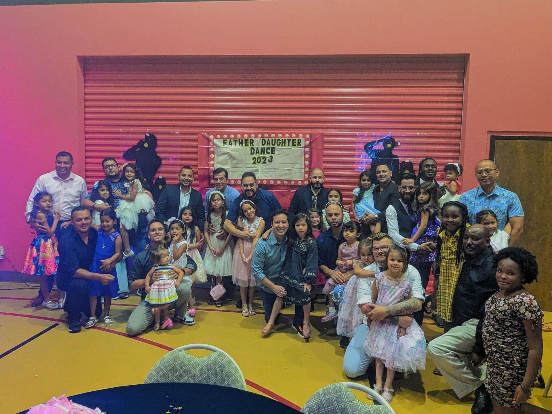 Group photo at event, people in formal attire, some with children, standing in front of sign on red wall.
