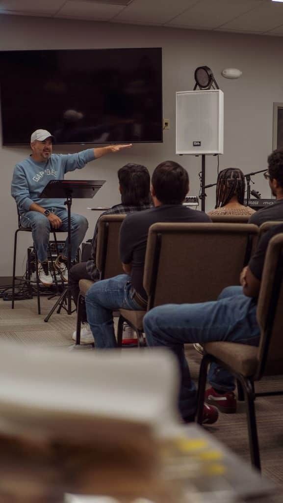 Man speaking to audience, gesturing with arm. Indoor setting, chairs, microphone, speaker, and TV visible.