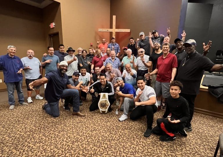Group of men posing with a championship belt, inside a building with a cross. Many are smiling and gesturing.