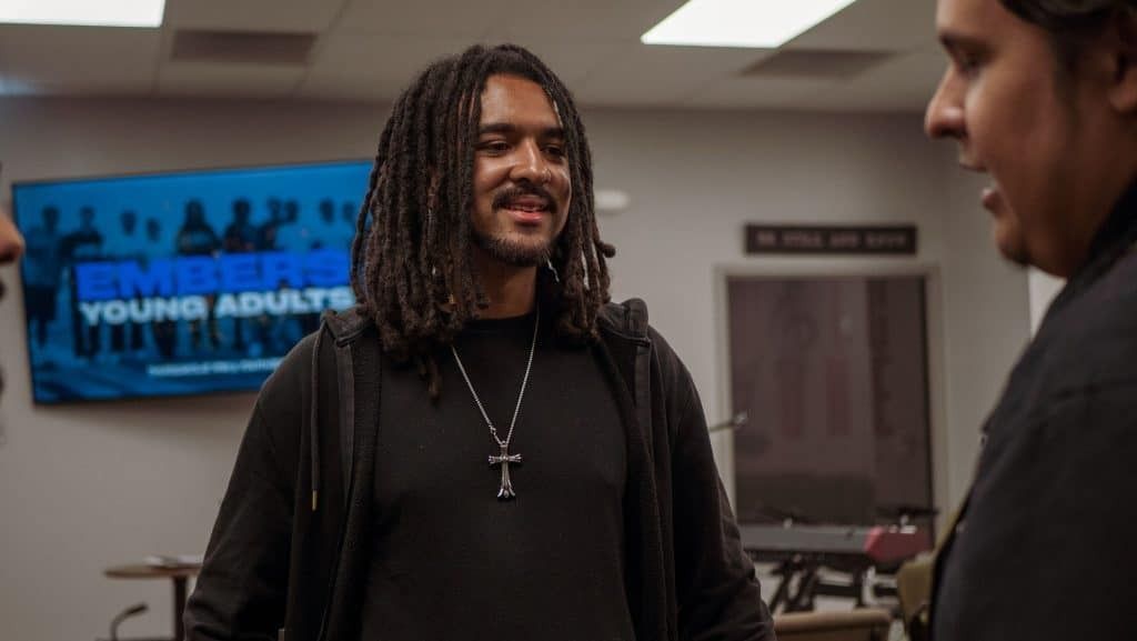 Man with dreadlocks and cross necklace smiles, talking to someone in a room.