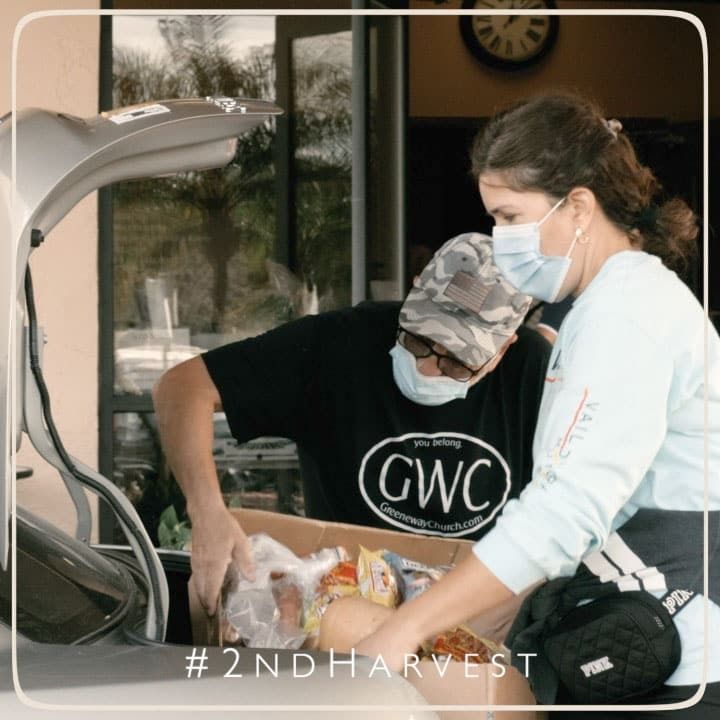 Volunteers loading groceries from box into a car trunk. They wear masks. Building in the background.
