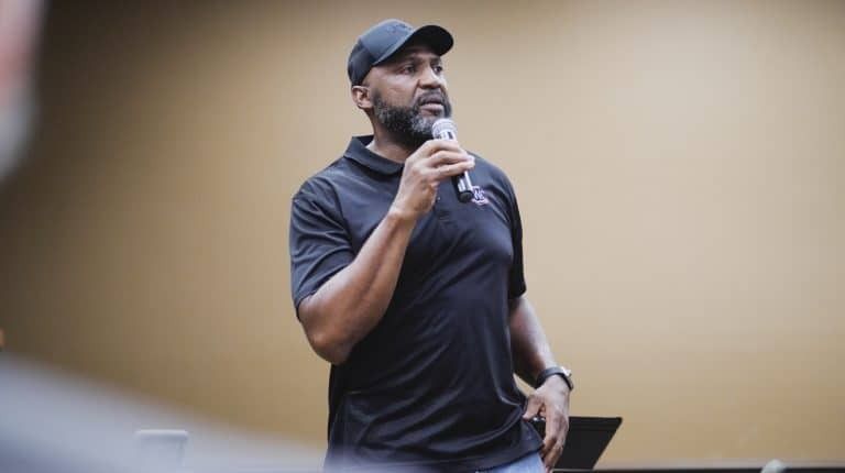 Man in black shirt and cap speaks into a microphone. Light-colored wall in background.