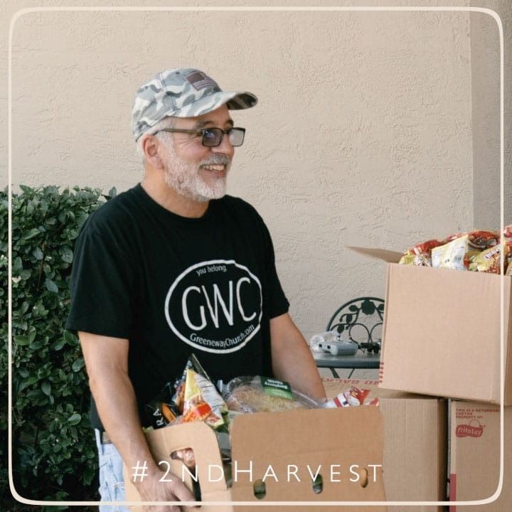 Man in black t-shirt and camouflage hat smiles while packing food boxes outside.