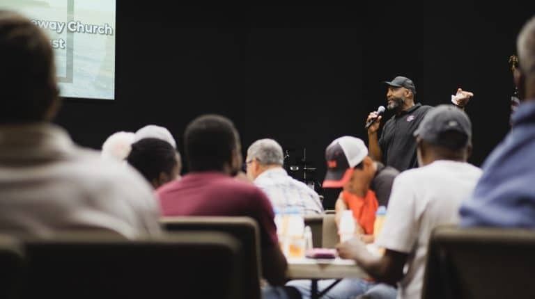 Man speaking into a microphone on stage, addressing a seated audience in a dimly lit auditorium.