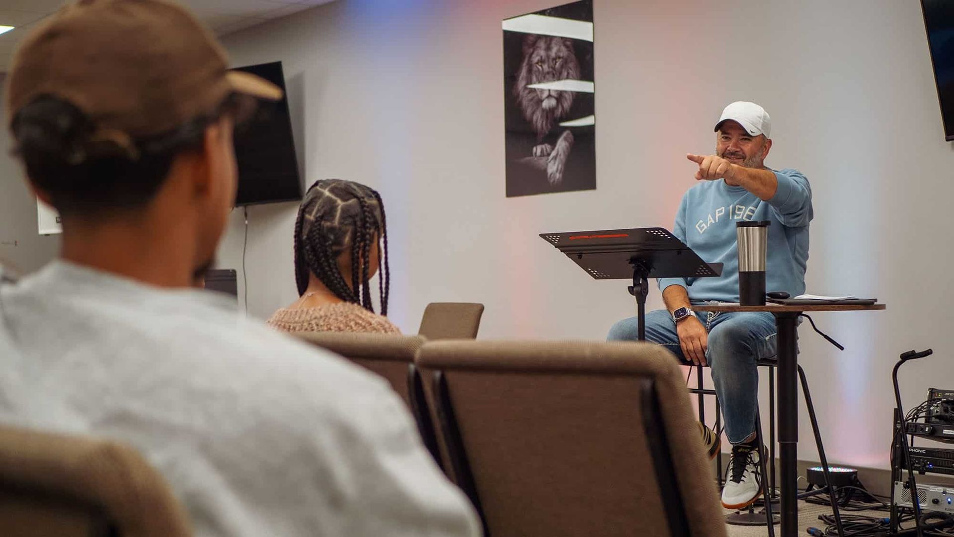 Man speaking from a podium to a small audience in a room. He points.