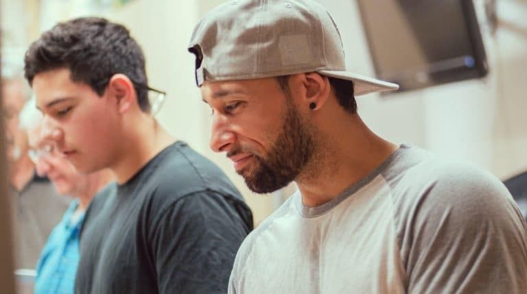 Man in baseball cap and light-colored shirt looks down, another man beside him, and a partial view of an older person.