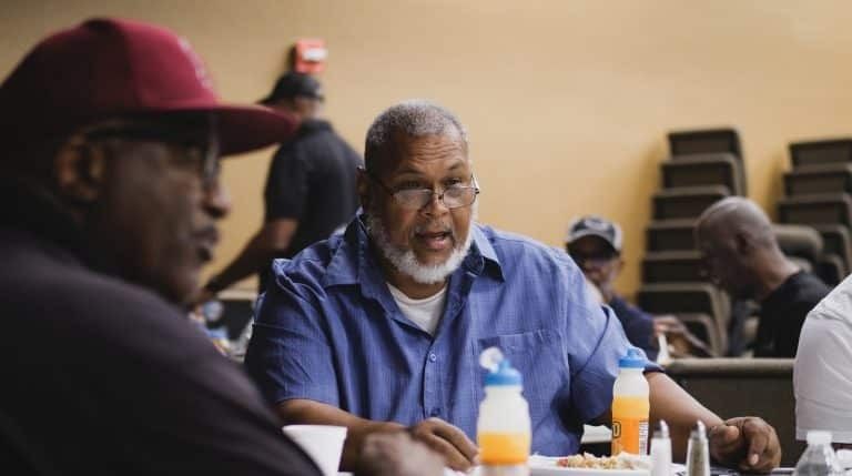 Men at a table, some wearing hats, having a discussion. Beverages and food are present in a large room.