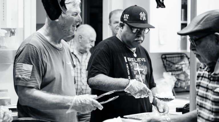 Men preparing food in a kitchen, wearing gloves, one in a Yankees cap, others observing.