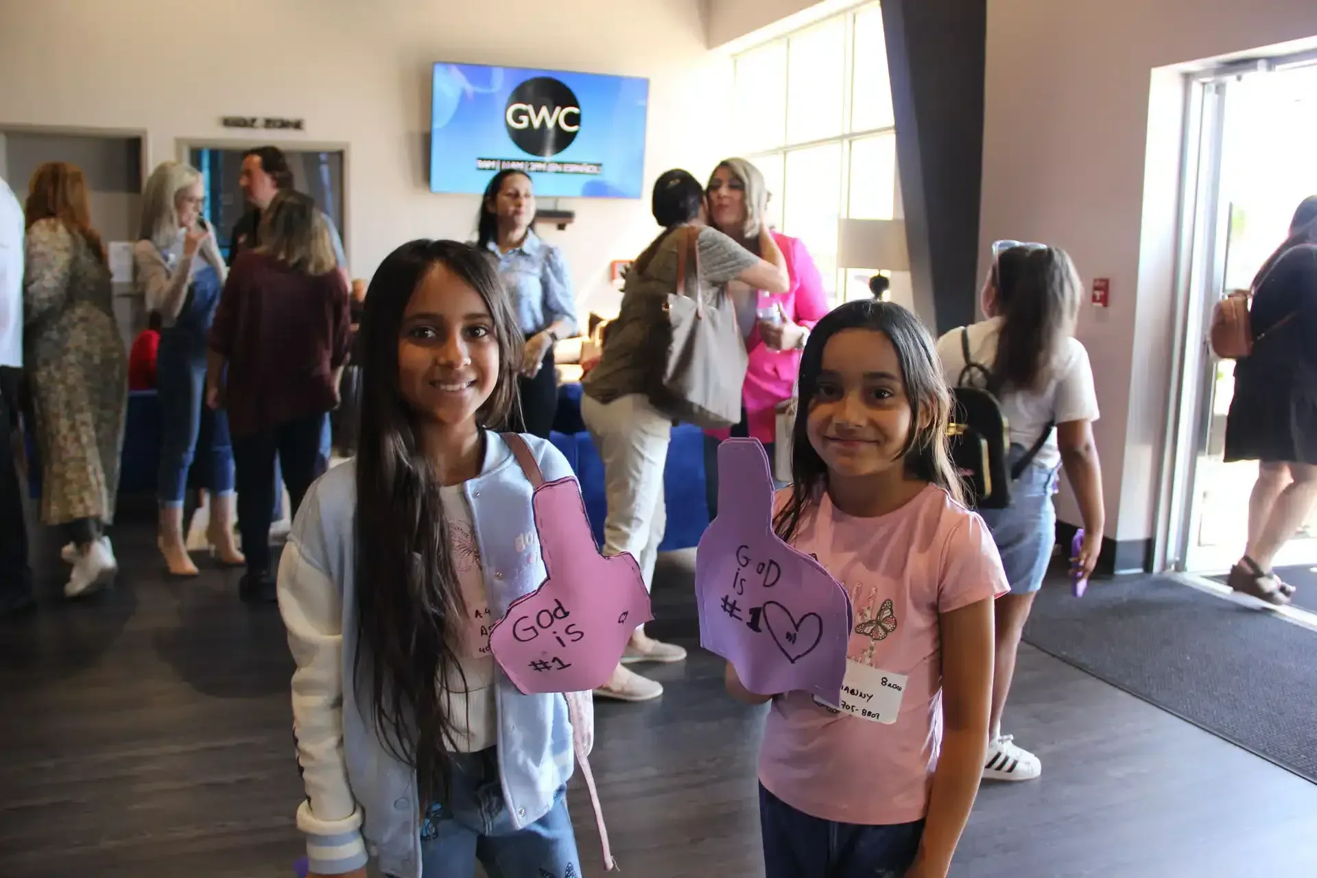 Two girls hold up paper thumbs-up signs at a gathering, other people in background.