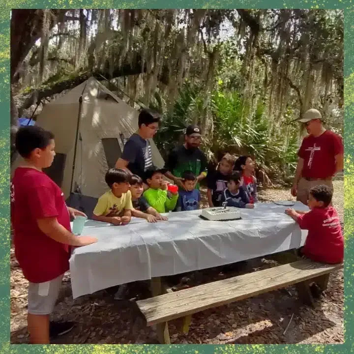 Group of people around a picnic table with food, a tent, and trees in a campsite setting.