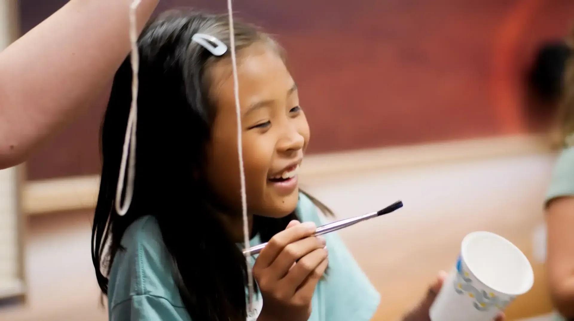 Smiling child holding a paintbrush and paper cup, with a string hanging in front of her.