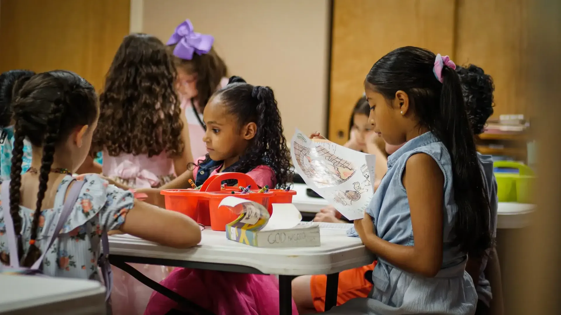 Children gathered around a table reading and working on projects in a brightly lit room.