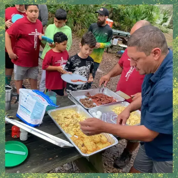 Group serving food from trays at a picnic table, other people watching.