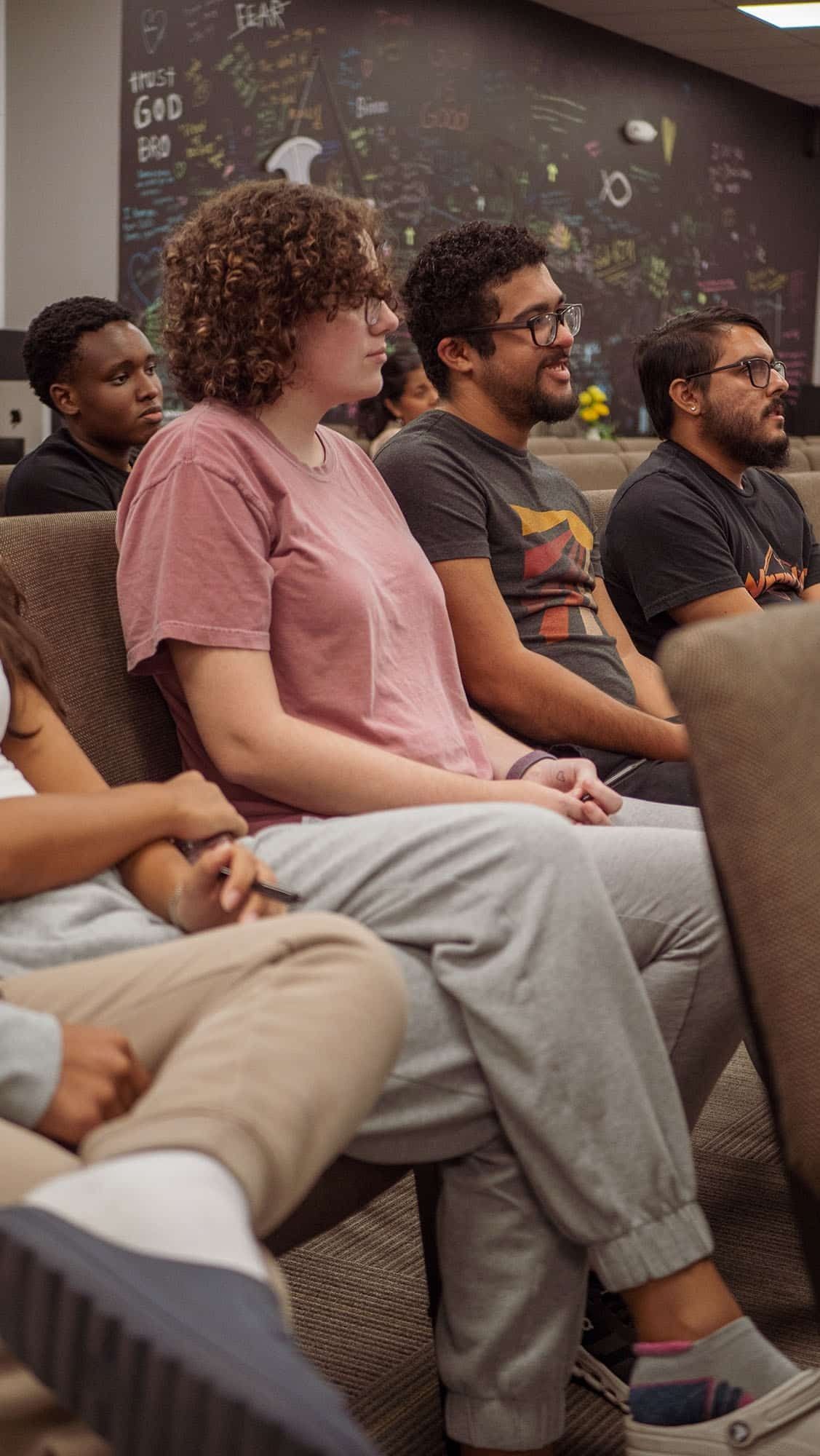People seated on chairs, listening in an auditorium; one wearing a pink shirt, others wearing t-shirts.