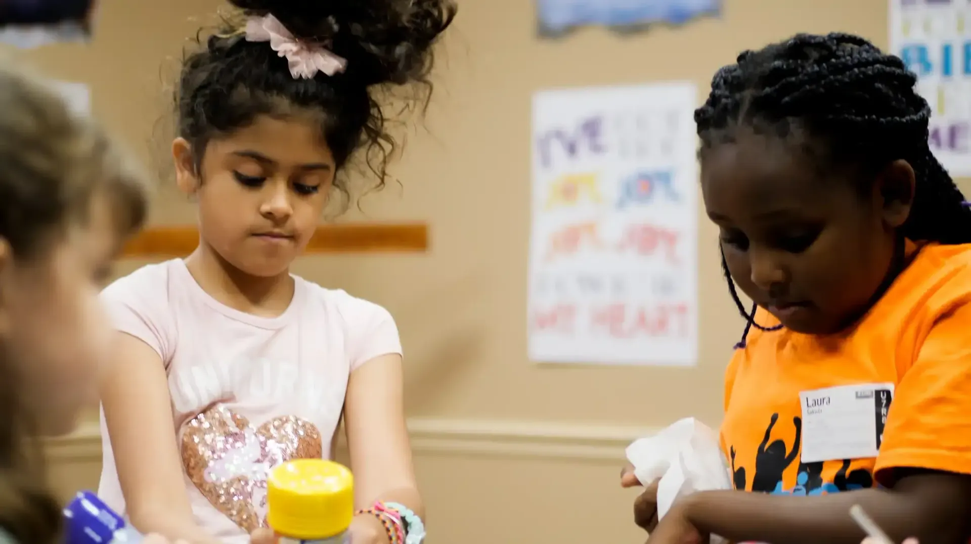 Two children focused on a craft project at a table. One wears an orange shirt, the other a pink shirt.