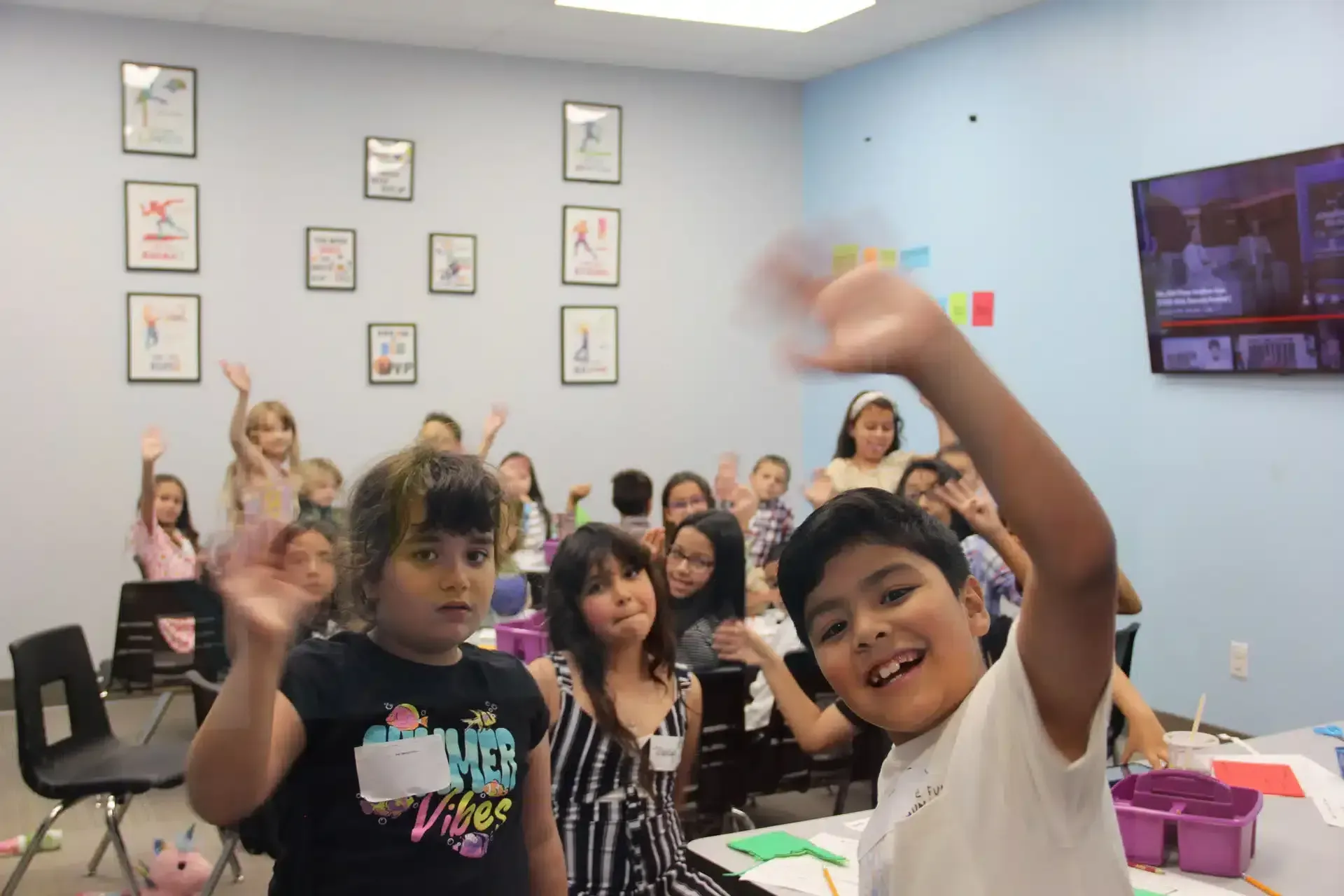 Children in a classroom waving with smiles; art on the walls.