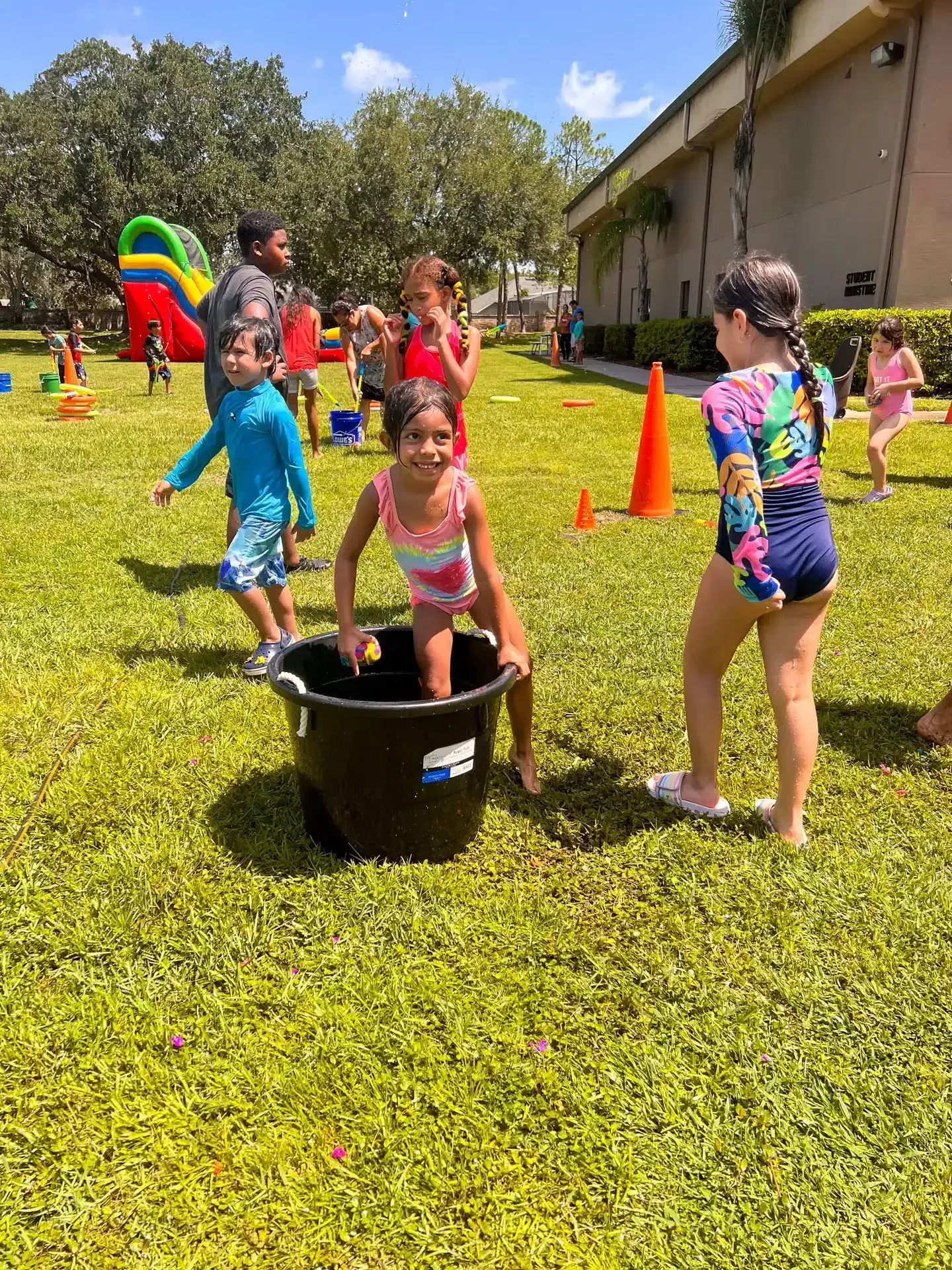 Children playing water games on a grassy lawn with a bounce house in the background.
