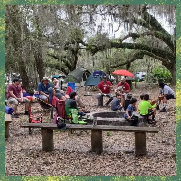 People gathered around a campfire at a campsite under trees with Spanish moss.