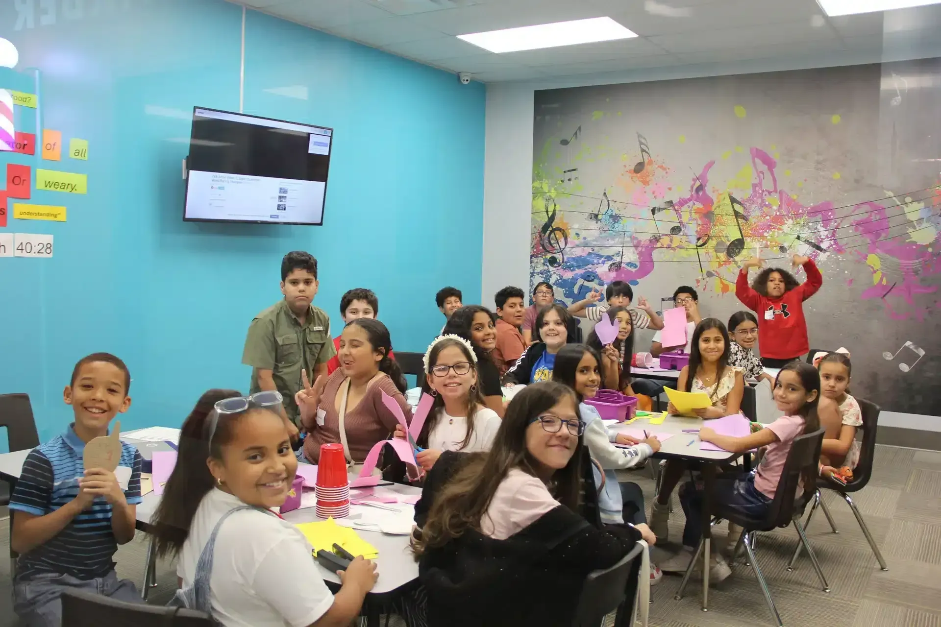 Children in a classroom making crafts; a few hold up their creations. Turquoise walls, colorful mural.