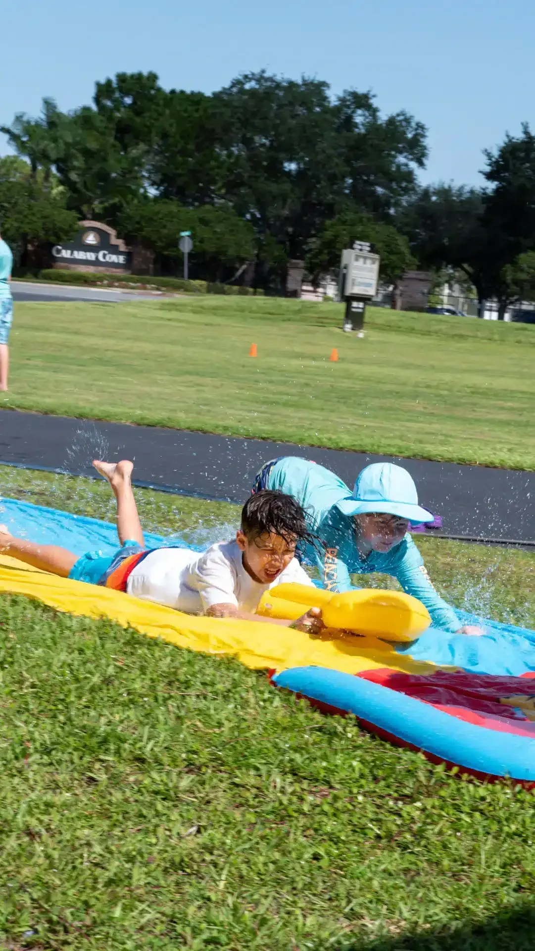 Two children slide on a water slide on a grassy field; one pushes the other.