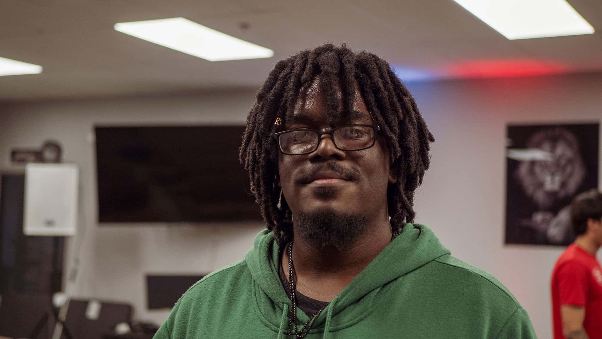Man with dreadlocks and glasses wearing a green hoodie, smiling, in a room with a large screen and artwork.