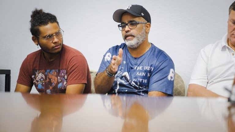 Three men seated at a table. Man in the center speaks, gesturing with hands; man on the left listens intently.
