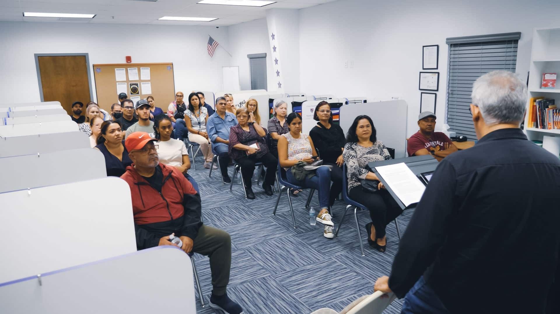 Audience listening to a speaker in a classroom. People sit in chairs, facing the man in the front.