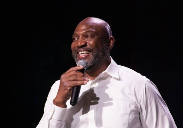 Man with shaved head and beard holding a microphone, smiling, against a black background.
