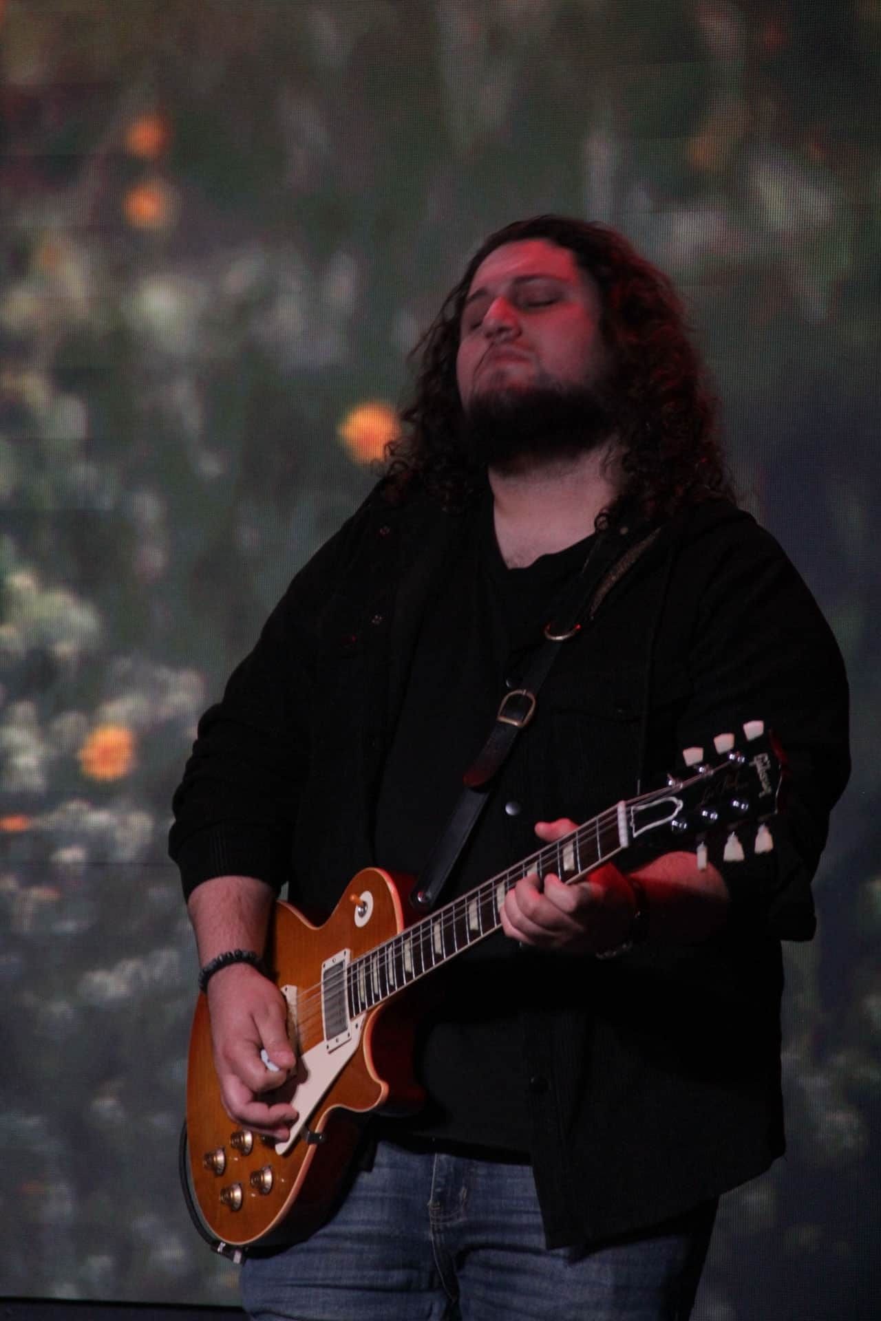 Guitarist with long hair playing a sunburst electric guitar, wearing black shirt and blue jeans, performing onstage.