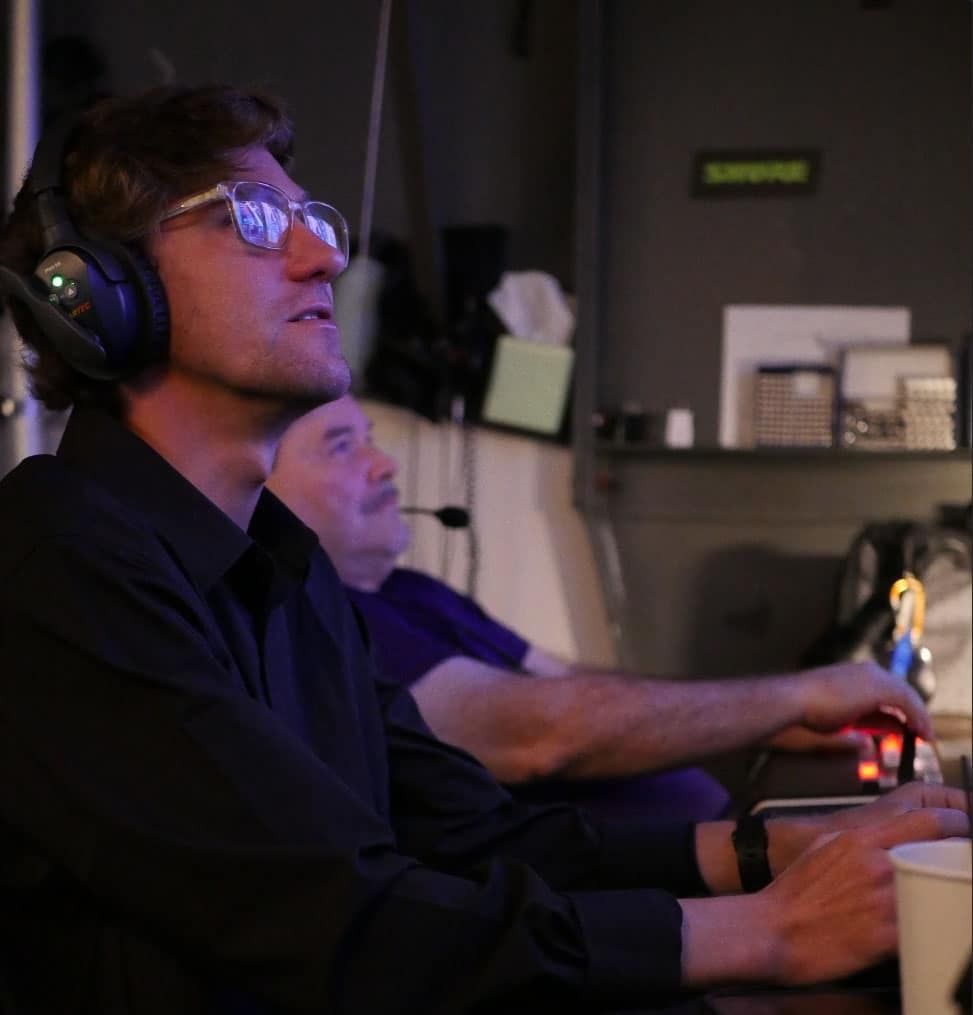 Man wearing headset and glasses, looking upward, seated at a computer, possibly in a studio setting.