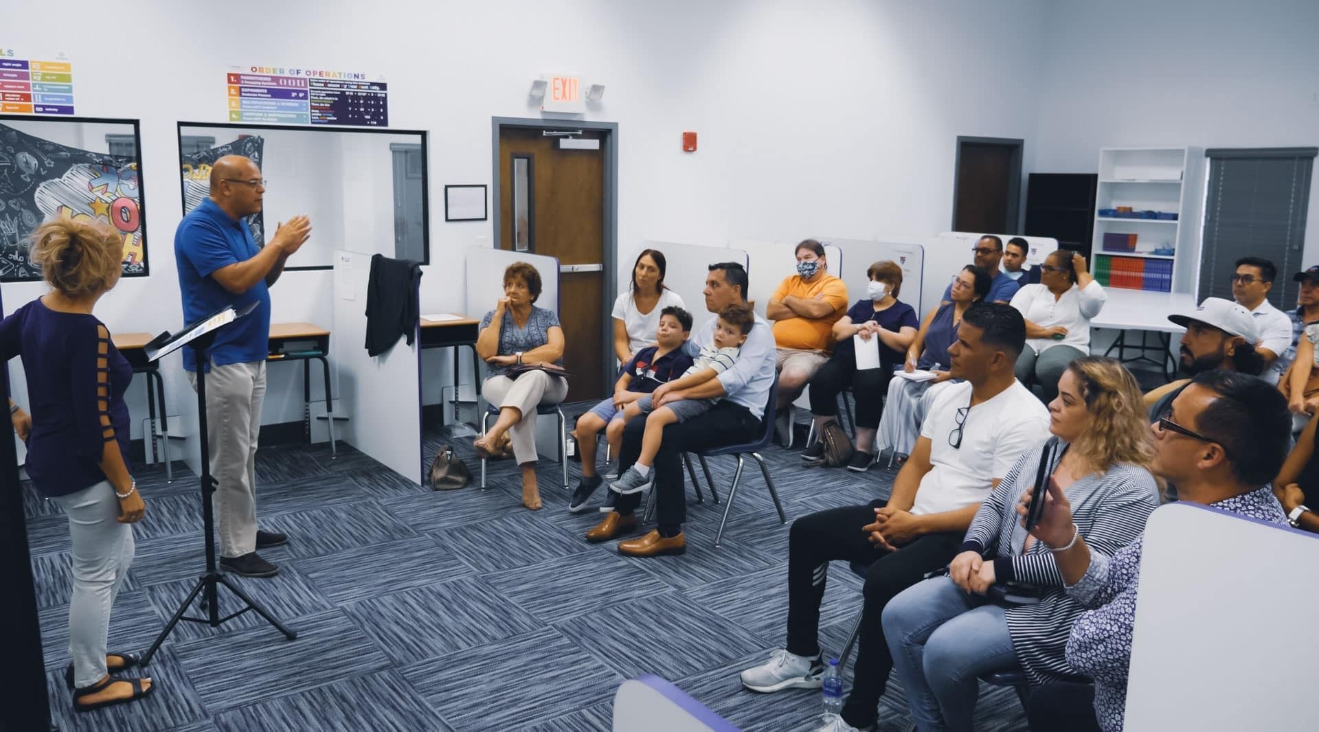 A man speaks to a seated audience in a room. A child stands on the side.