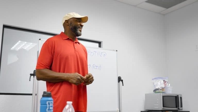 Man in red shirt and tan cap speaking, whiteboard in background.