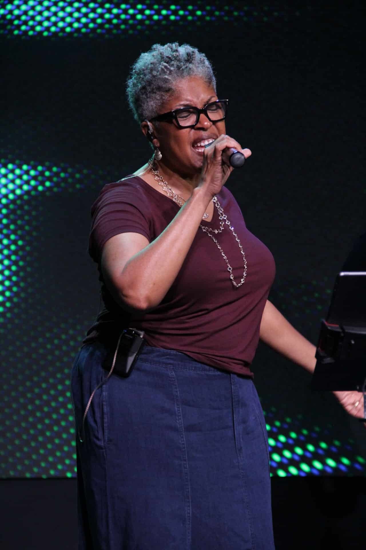 Woman singing into a microphone on stage, wearing glasses and a maroon shirt.