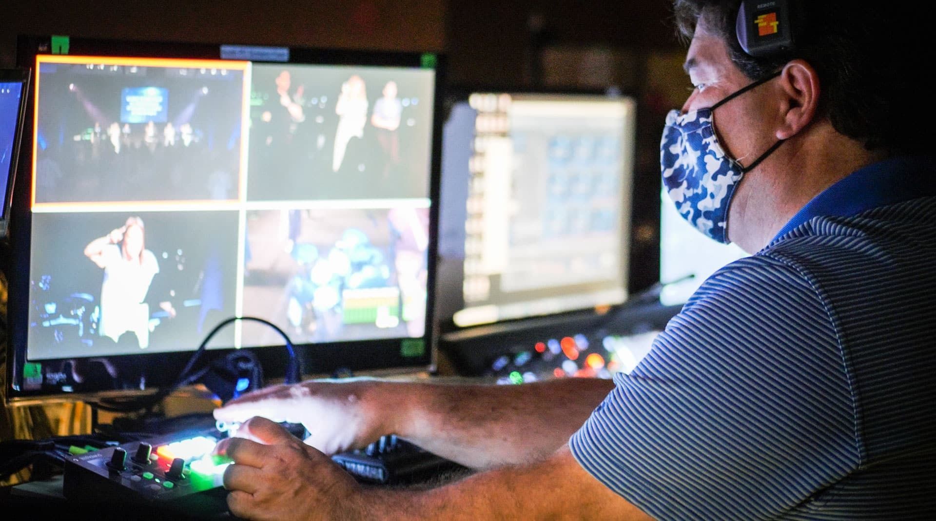 Person wearing a mask and headset operating video equipment, monitoring live footage on multiple screens.