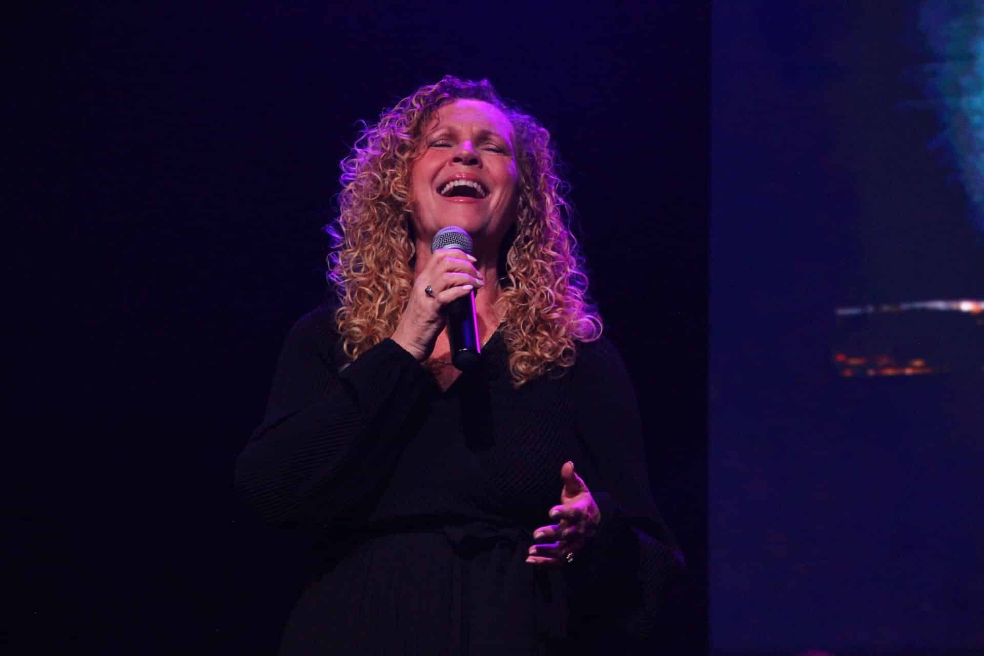 Woman singing into a microphone on stage, smiling and looking up, with a dark background.