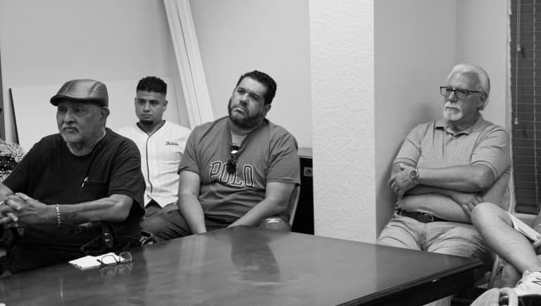 A group of men sitting at a table indoors. Some look towards a speaker.
