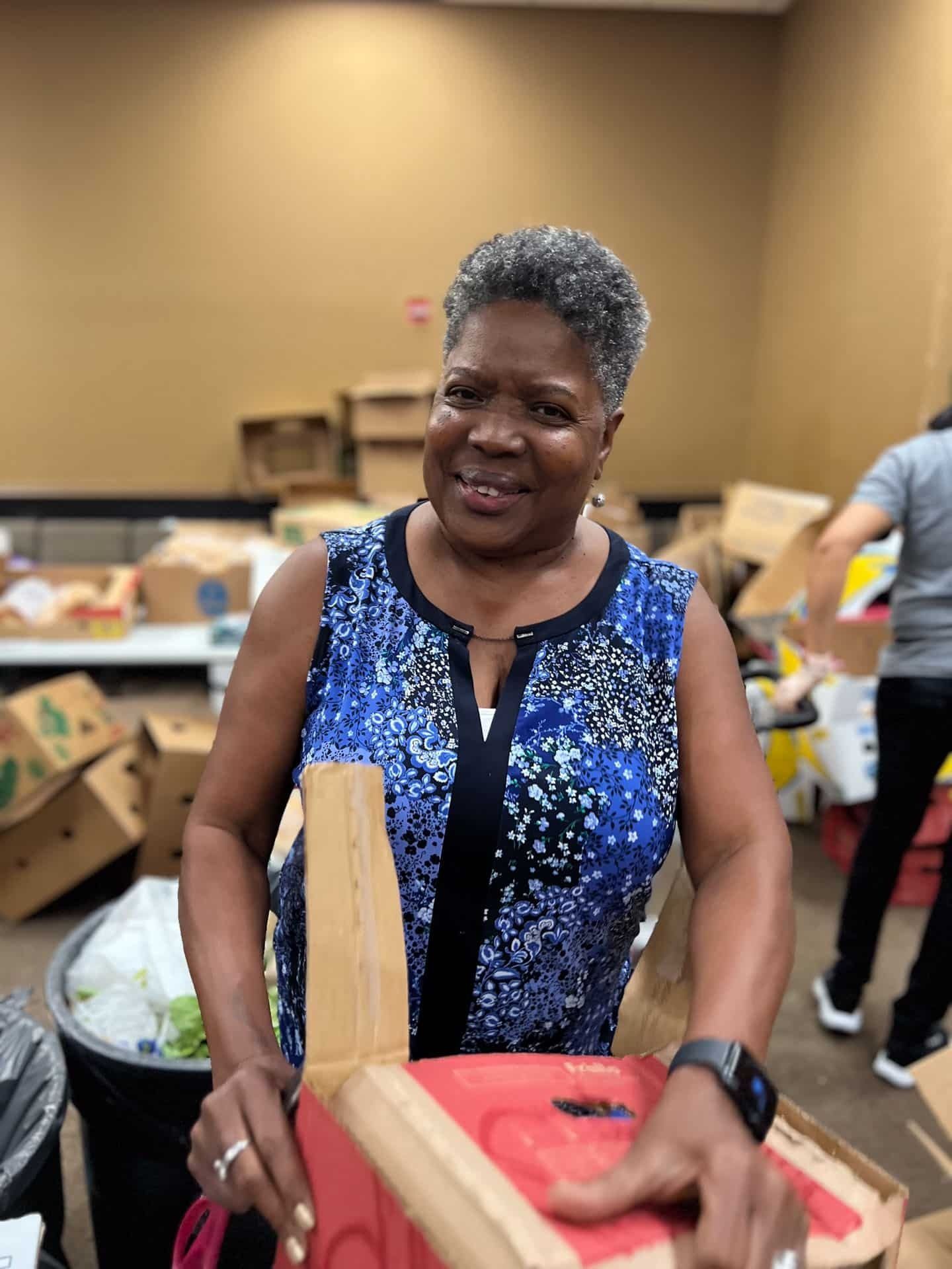 Woman in blue floral top smiling, holding a red and tan object, in a room with boxes.