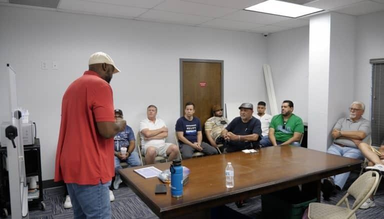 Man in red shirt speaks to group seated at a table in a room.