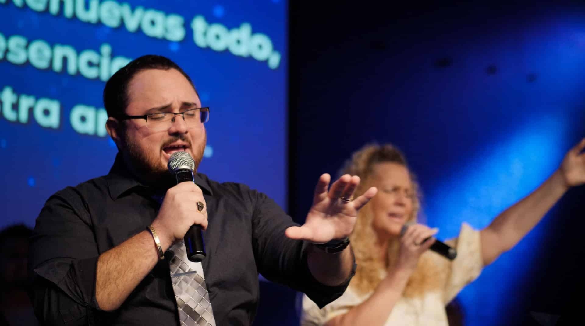 Man singing into a microphone, hands outstretched. Woman sings with arm raised; blue lighting in background.
